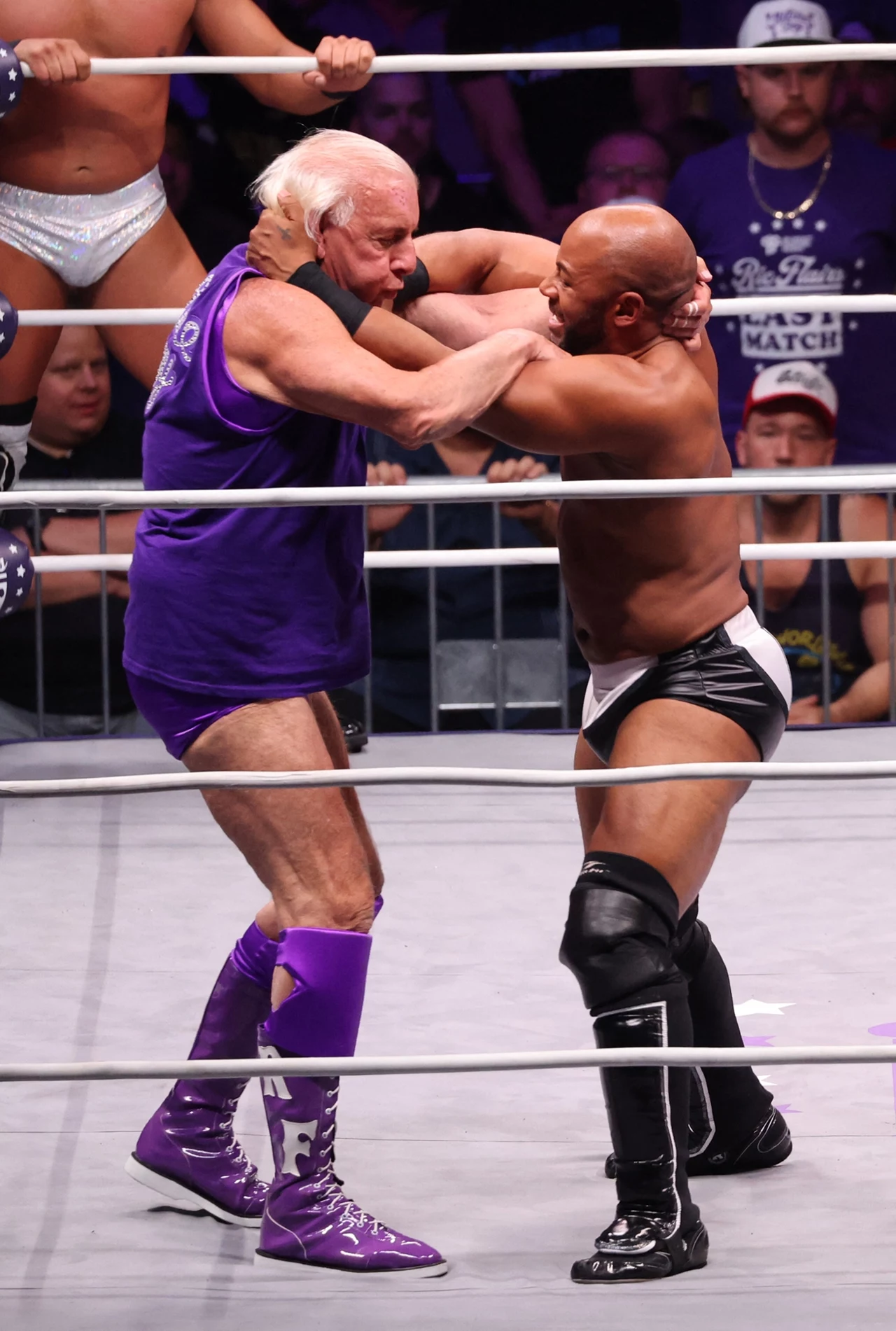 Ric Flair (po lewej) Dwóch mężczyzn w ringu wrestlingowym trzyma się za szyje i ramiona, walcząc ze sobą, wokół ring otoczony widzami, jeden zawodnik ubrany jest w fioletowy strój, drugi w czarno-białe spodenki.