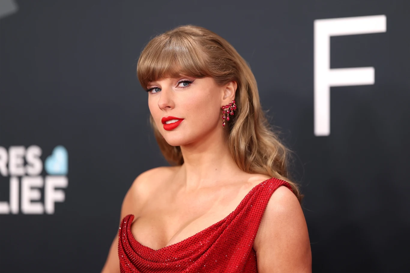 Taylor Swift wydała "folklore" 5 lat temu Kobieta z długimi blond włosami pozuje w eleganckiej, czerwonej sukni z dekoltem, z makijażem podkreślającym usta czerwonym kolorem, w tle rozmyta powierzchnia z dużą białą literą F.