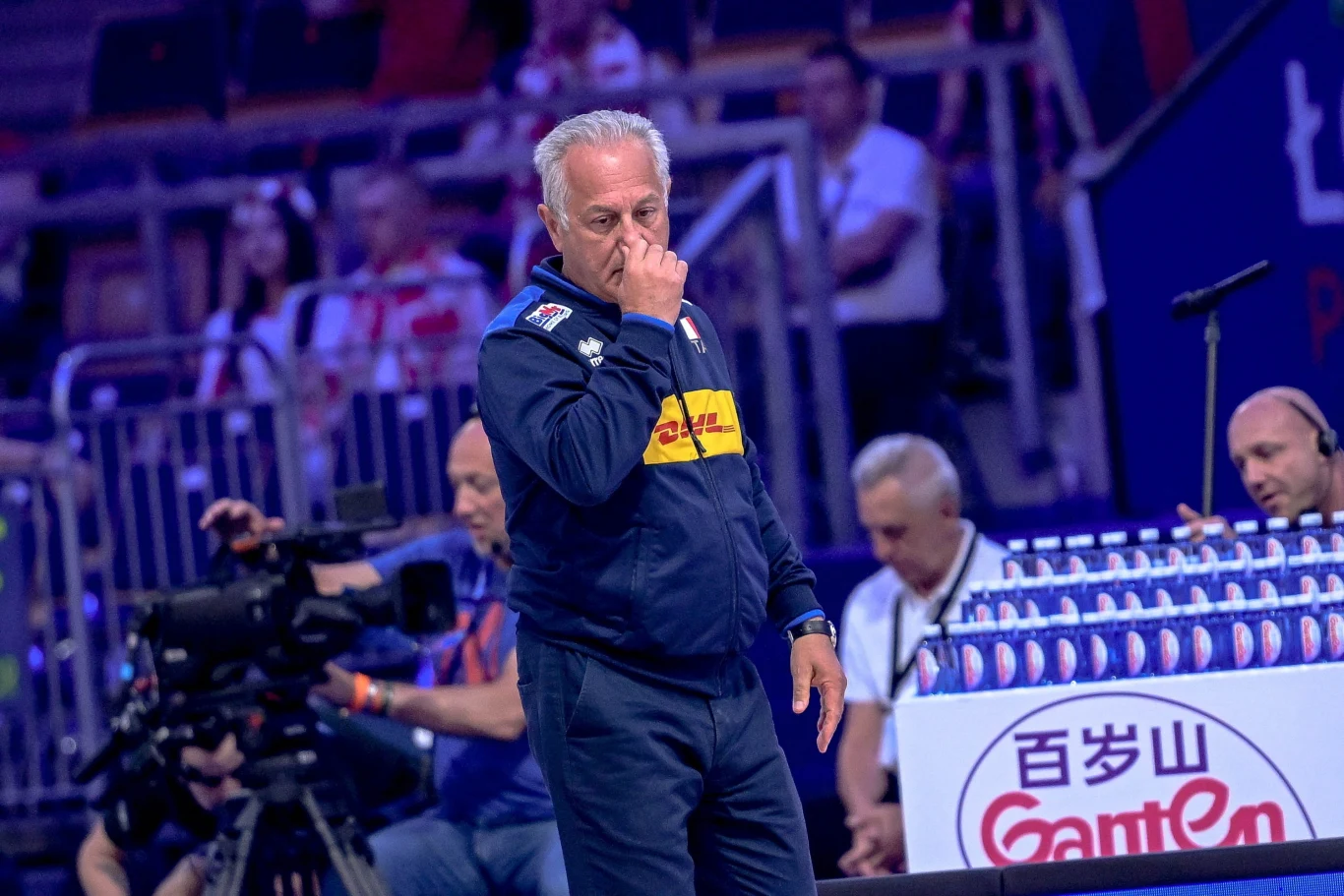 Trener w sportowym stroju myśli podczas meczu siatkówki, w tle widoczne trybuny z kibicami oraz kamera telewizyjna i stanowisko komentatorów