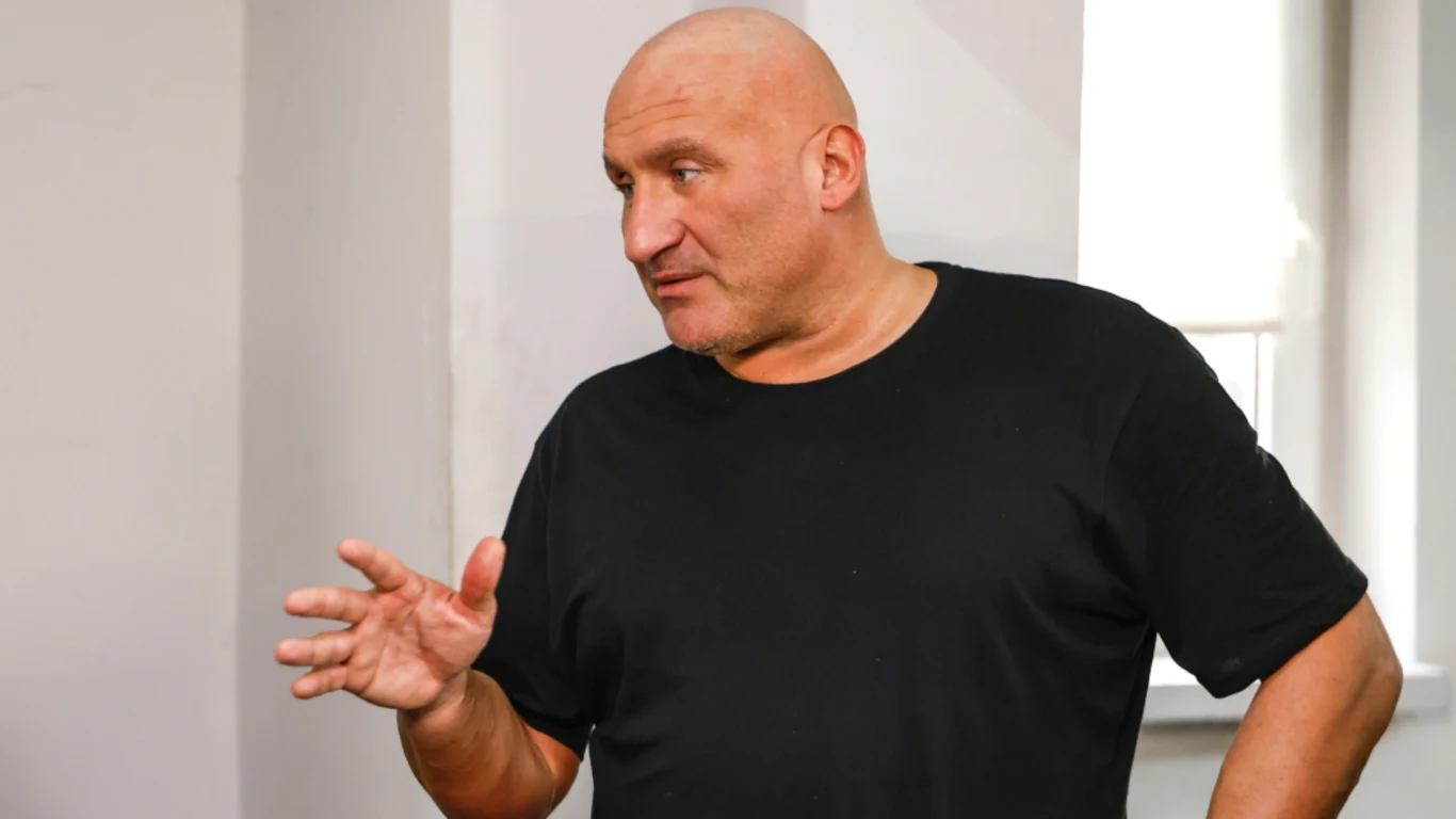 Łysy mężczyzna w czarnej koszulce gestykuluje ręką, stojąc w jasnym pomieszczeniu obok okna.