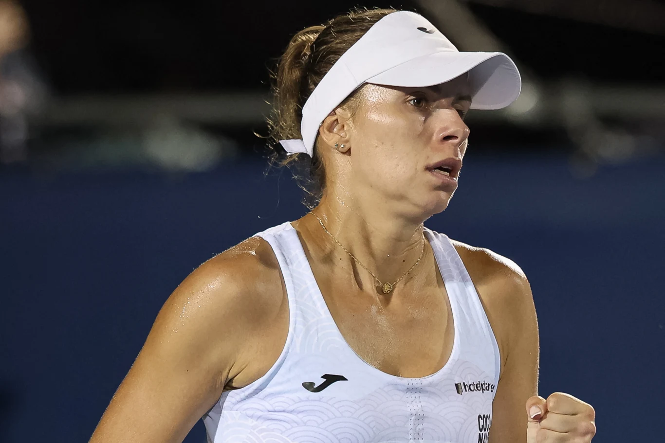 Kobieta w sportowej odzieży uczestnicząca w meczu tenisowym, z widocznym wyrazem determinacji i zaangażowania, trzyma zaciśniętą pięść, na głowie ma biały daszek sportowy, a tło jest rozmyte.