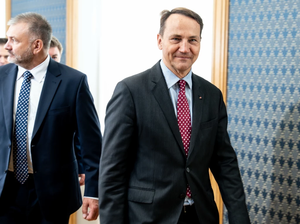 Radosław Sikorski i Waldemar Żurek podczas ogłaszania przez Donalda Tuska rekonstrukcji rządu Dwaj mężczyźni w garniturach idą korytarzem, jeden z nich uśmiechnięty, drugi ma poważniejszy wyraz twarzy, w tle widoczne są częściowo inne osoby i wzorzysty niebieski panel ścienny.