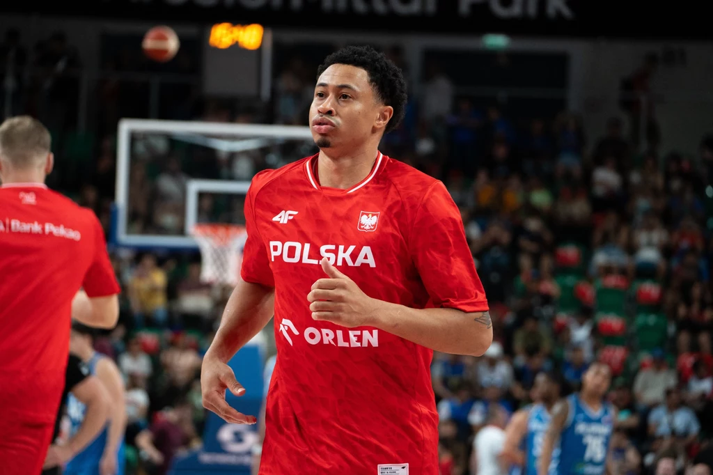 A.J. Slaughter Koszykarz w czerwonym stroju reprezentacji Polski podczas rozgrzewki na hali sportowej, w tle widoczni inni zawodnicy oraz kibice siedzący na trybunach.