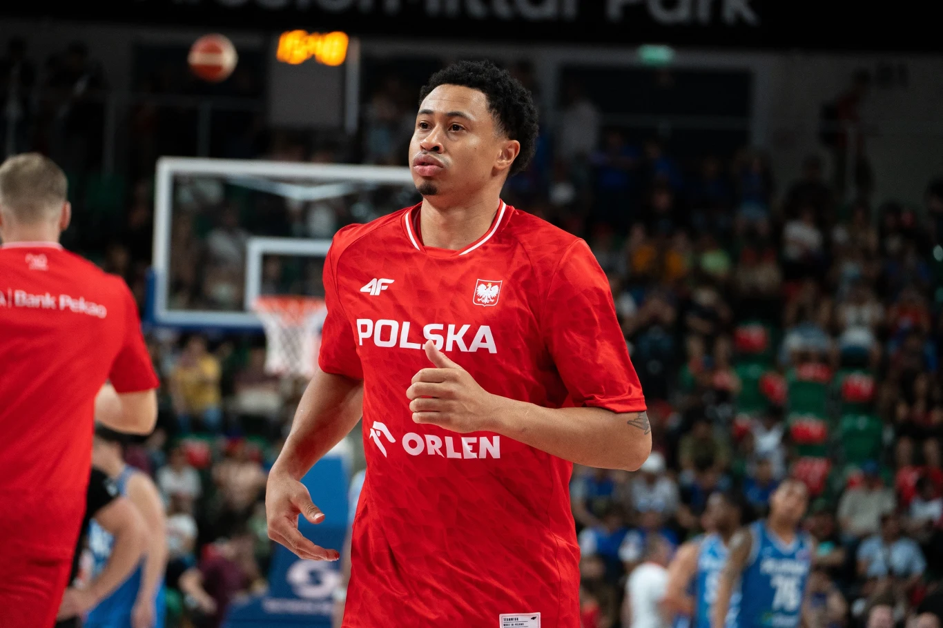 Koszykarz w czerwonym stroju reprezentacji Polski podczas rozgrzewki na hali sportowej, w tle widoczni inni zawodnicy oraz kibice siedzący na trybunach.