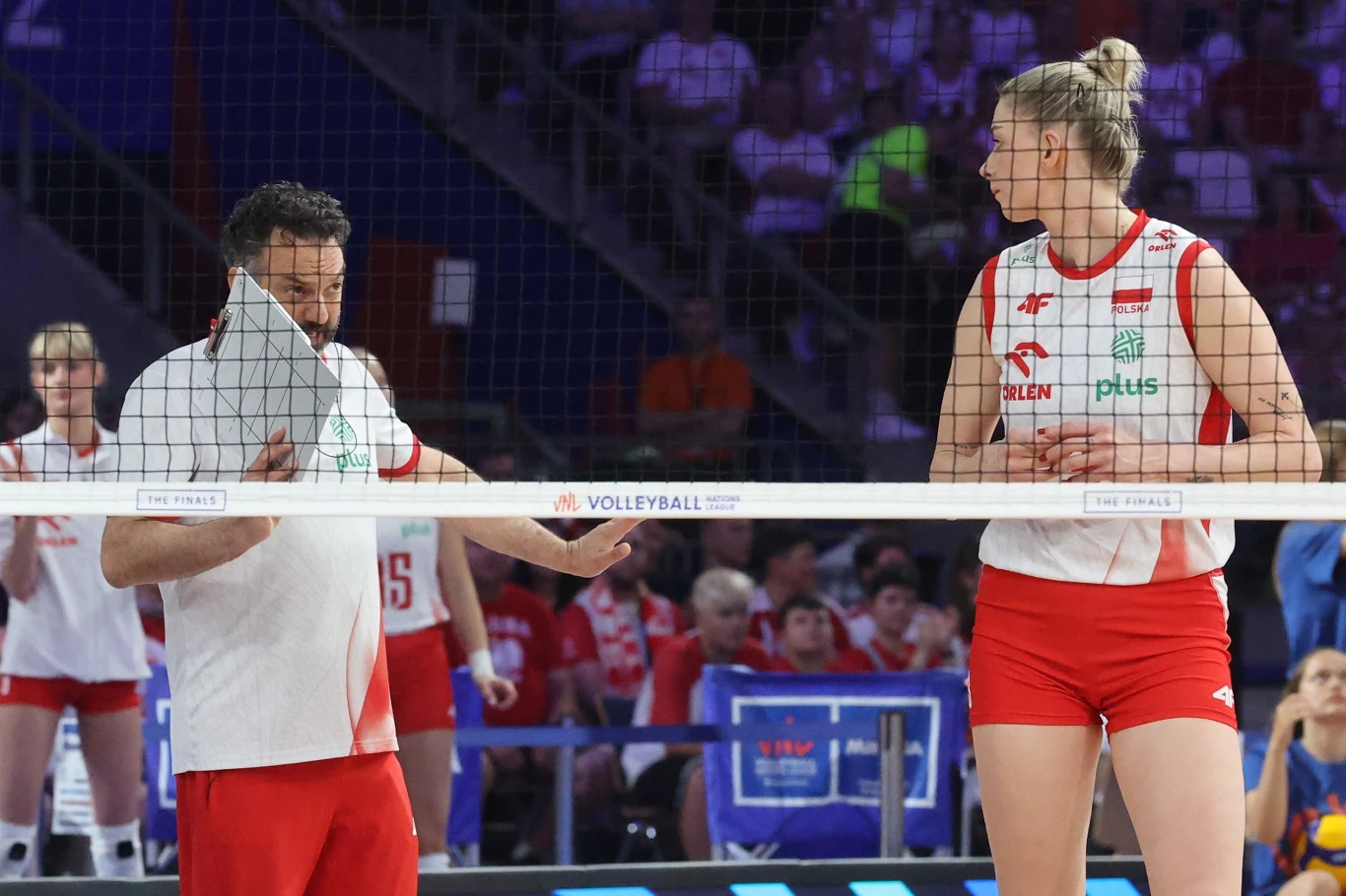 Trener rozmawia z siatkarką drużyny narodowej Polski podczas meczu, oboje znajdują się po przeciwnych stronach siatki, w tle widownia kibiców.