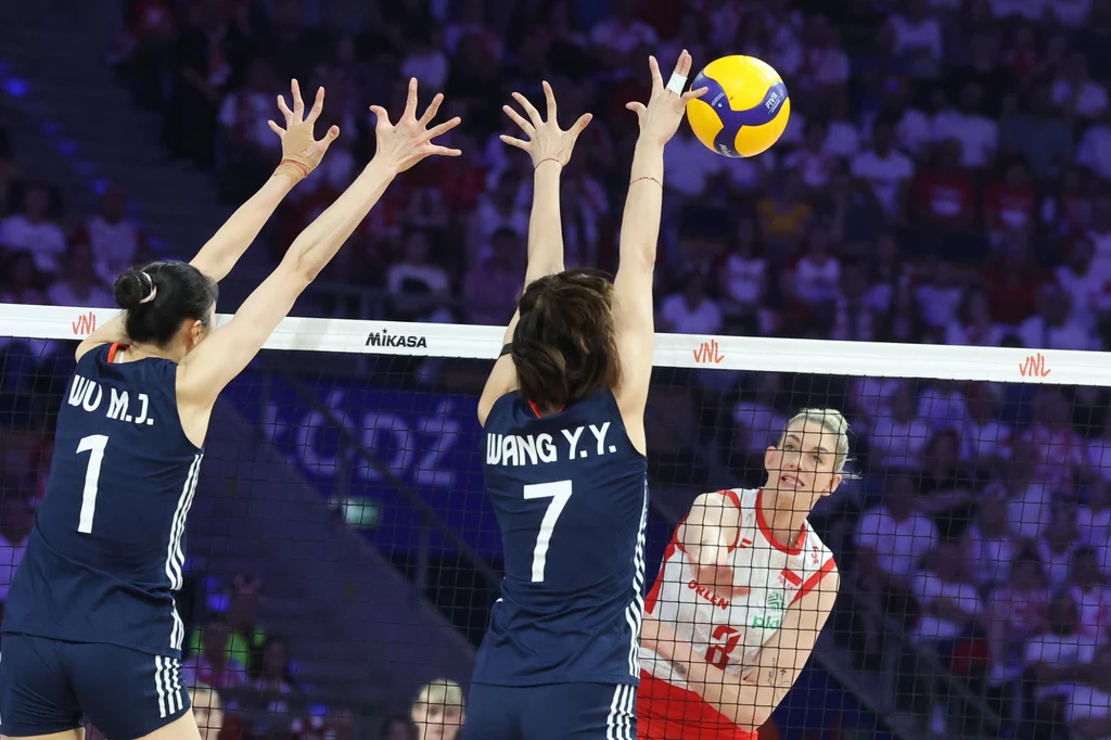 Kadr z meczu Polska - Chiny Siatkarka w biało-czerwonym stroju atakuje piłkę nad siatką, podczas gdy dwie zawodniczki drużyny przeciwnej w granatowych strojach próbują zablokować uderzenie, uniesione ręce po obu stronach siatki, dynamiczna atmosfera hali sportowej, liczna publicz...