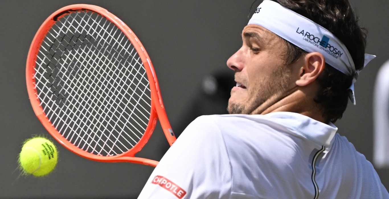 Taylor Fritz Zbliżenie na tenisistę w białej koszulce z opaską na głowie, skupionego na odbiciu piłki rakietą sportową. W tle rozmyte elementy kortu.