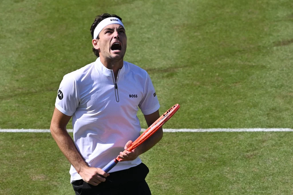 Taylor Fritz Zawodnik tenisowy w białej koszulce i opasce na głowie trzyma w ręce rakietę, wyrażając silne emocje, podczas gry na trawiastym korcie.
