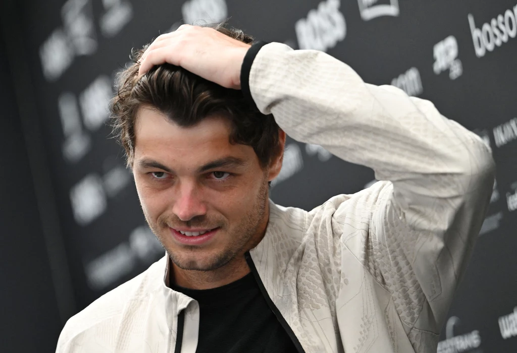 Taylor Fritz Mężczyzna w jasnej kurtce poprawiający włosy na tle czarnej ścianki z logo i napisami.