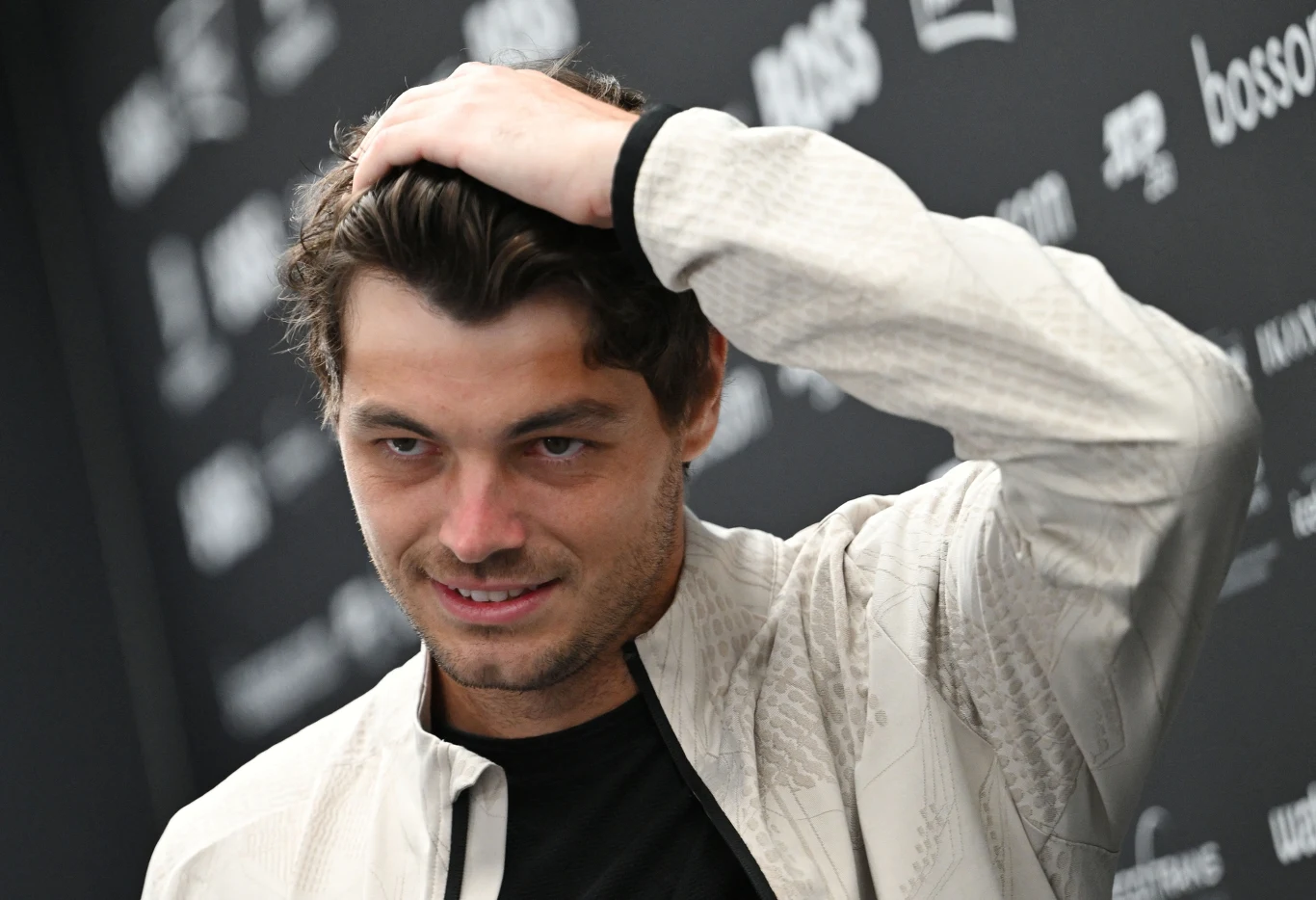 Taylor Fritz Mężczyzna w jasnej kurtce poprawiający włosy na tle czarnej ścianki z logo i napisami.