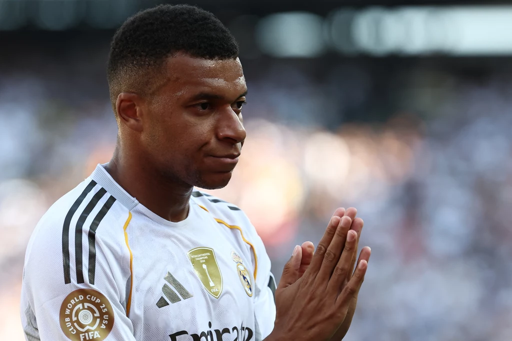 Kylian Mbappe Młody mężczyzna w białej koszulce drużyny piłkarskiej Realu Madryt stoi ze złożonymi dłońmi, na tle rozmytej widowni stadionu, z poważnym wyrazem twarzy.