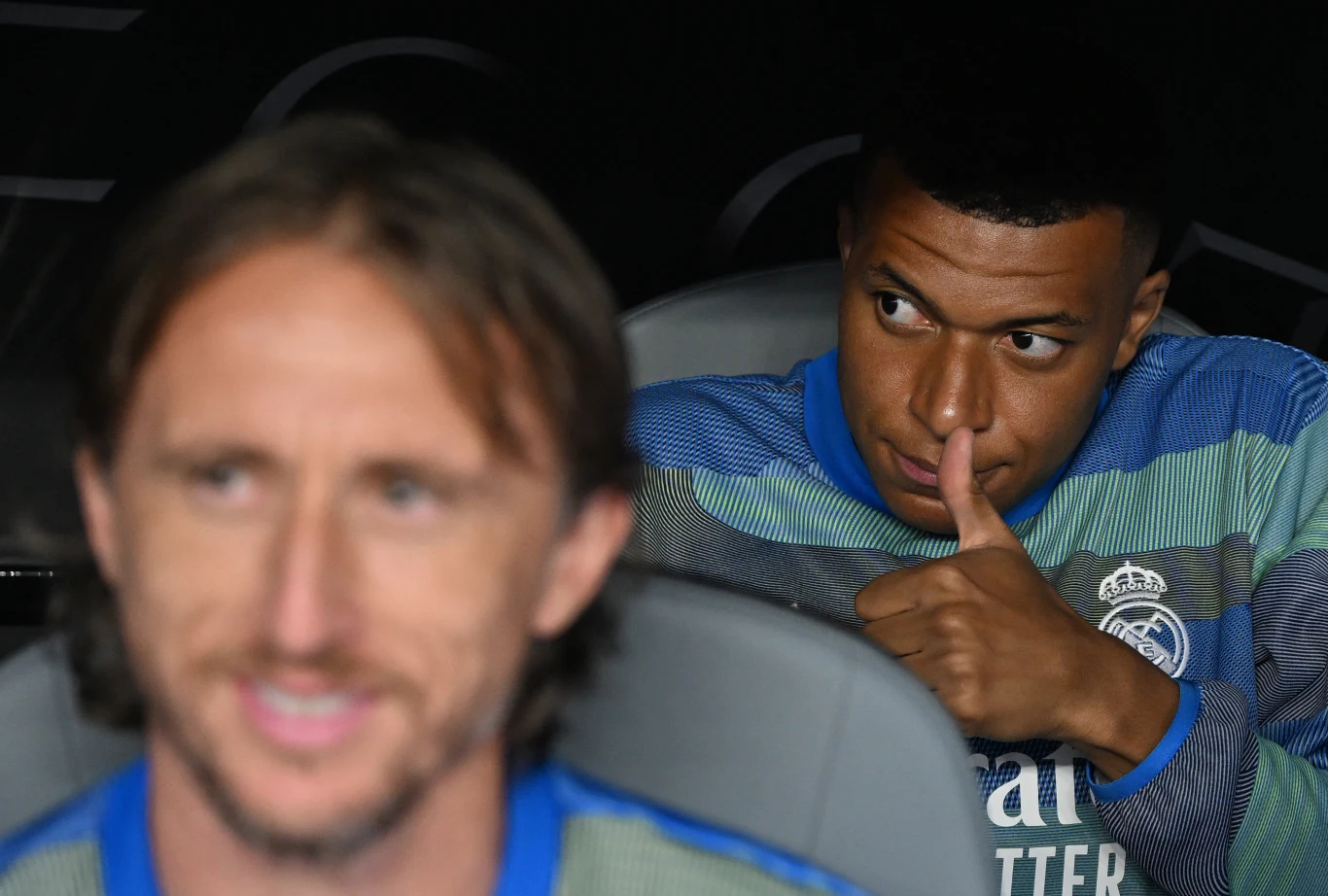 Dwóch piłkarzy w sportowych strojach drużyny Real Madryt na ławce rezerwowych, jeden z nich pokazuje gest kciuka w górę i uśmiecha się, drugi znajduje się na pierwszym planie z rozmazaną twarzą.