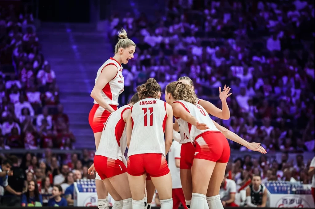 Liga Narodów siatkarek. Reprezentantki Polski w trakcie meczu z Chinami Grupa zawodniczek polskiej drużyny siatkarskiej w białych i czerwonych strojach zebrania w kółku na boisku, wokół nich kibice na trybunach.