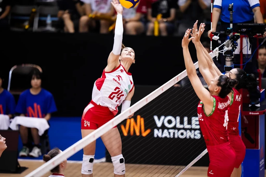 Liga Narodów siatkarek. Paulina Damaske atakuje Zawodniczka w białoczerwonym stroju wykonuje atak w siatkówce, unosząc wysoko rękę, podczas gdy dwie przeciwniczki w czerwonych strojach próbują zablokować piłkę przy siatce, w tle widoczne logo turnieju i widzowie.