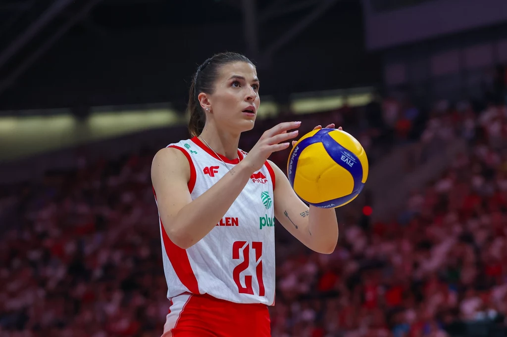 Alicja Grabka Zawodniczka siatkarskiej reprezentacji Polski w narodowym stroju, trzymająca piłkę do siatkówki i przygotowująca się do wykonania serwisu na hali sportowej, w tle rozmyta publiczność.