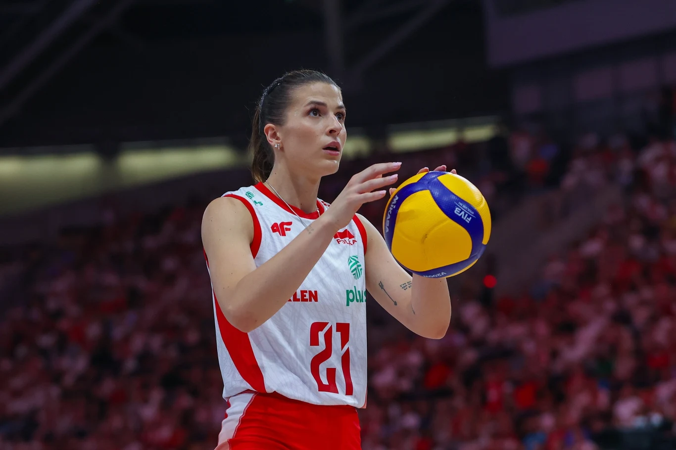 Alicja Grabka Zawodniczka siatkarskiej reprezentacji Polski w narodowym stroju, trzymająca piłkę do siatkówki i przygotowująca się do wykonania serwisu na hali sportowej, w tle rozmyta publiczność.