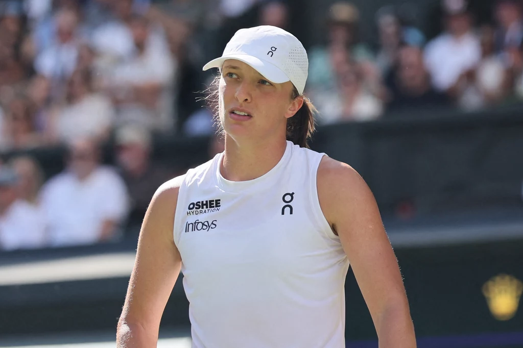 Iga Świątek Tenisistka w białym stroju sportowym i czapce, stojąca na korcie z poważnym wyrazem twarzy, w tle rozmyta publiczność.