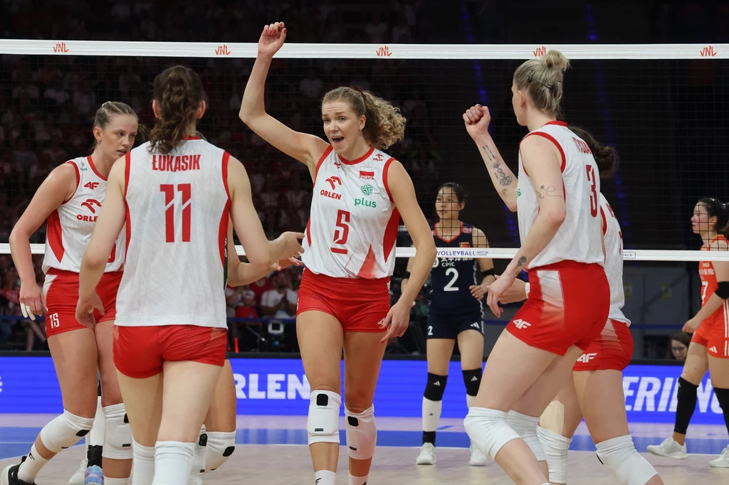 Zawodniczki polskiej reprezentacji siatkówki świętujące punkt podczas meczu, ubrane w biało-czerwone stroje z wyraźnymi nazwiskami i numerami, w tle siatka oraz rywalki, atmosfera intensywnej rywalizacji sportowej.