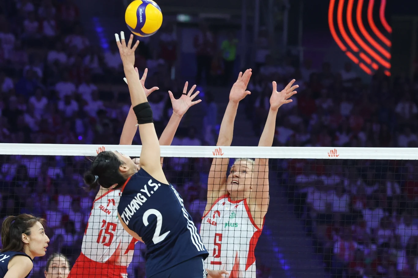 Blok Polek w meczu z Chinkami Dwóch siatkarek w białych strojach z czerwonymi akcentami próbuje zablokować atak zawodniczki w granatowej koszulce na tle pełnej widowni podczas meczu siatkówki.