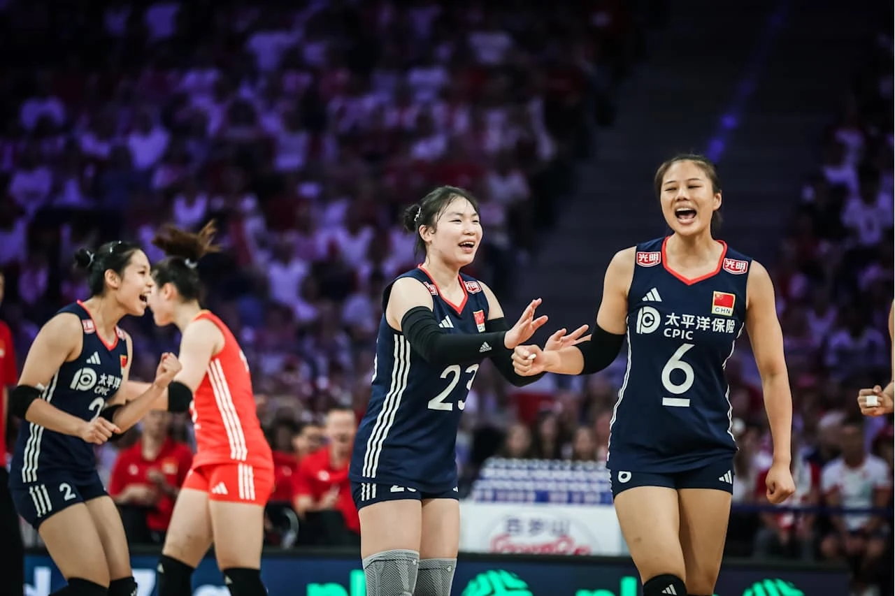 Reprezentantki Chin Zawodniczki drużyny siatkówki w ciemnych strojach świętujące podczas meczu w hali sportowej, w tle rozmyta publiczność oraz inne zawodniczki w strojach czerwonych.