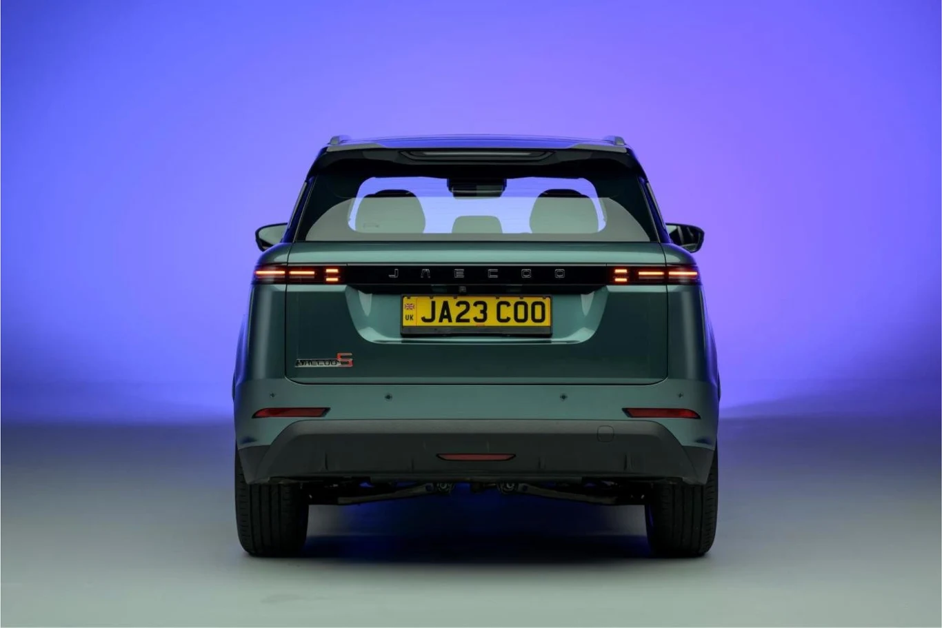 Widok tylnej części zielonego samochodu osobowego JAECOO, z widoczną tablicą rejestracyjną JA23 COO, na jednolitym fioletowo-niebieskim tle studyjnym, światła LED oraz delikatnie widoczne wnętrze pojazdu przez tylne szyby.