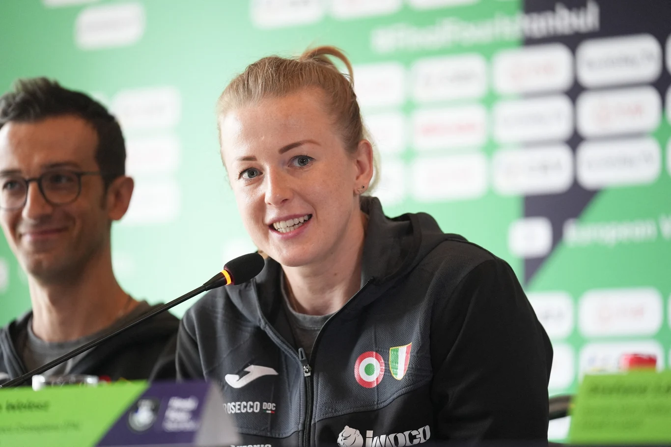 Joanna Wołosz Uśmiechnięta kobieta w sportowej bluzie z emblematami klubowymi siedząca przy mikrofonie podczas konferencji prasowej, w tle zarysowany baner z logo radia sportowego i nazwami sponsorów.