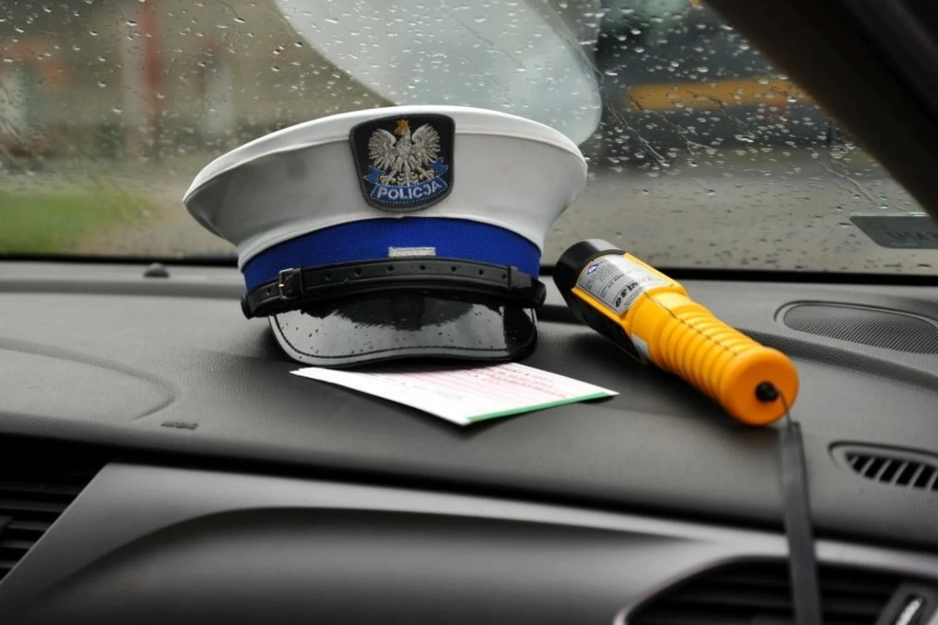 Czapka polskiej policji, bloczek mandatowy oraz alkotest leżące na desce rozdzielczej samochodu na tle deszczu padającego na szybę.