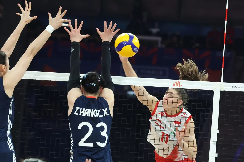Dwóch zawodników drużyny w granatowych strojach próbuje zablokować atakującego siatkarkę w białoczerwonym stroju uderzającą piłkę nad siatką, w dynamicznej akcji sportowej podczas meczu siatkówki.