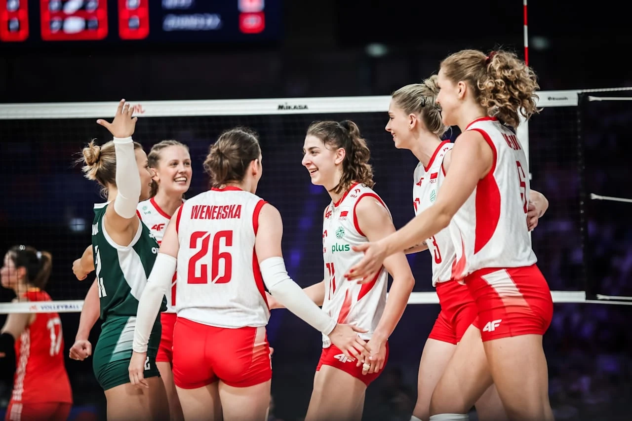 Liga Narodów siatkarek. Polskie siatkarki pokonały Chinki, teraz czas na Włoszki Grupa zawodniczek polskiej reprezentacji siatkówki w białych i czerwonych strojach cieszy się po zdobyciu punktu podczas meczu, dziewczyny są uśmiechnięte i okazują radość oraz sportową energię, w tle siatka i elementy hali sportowej.