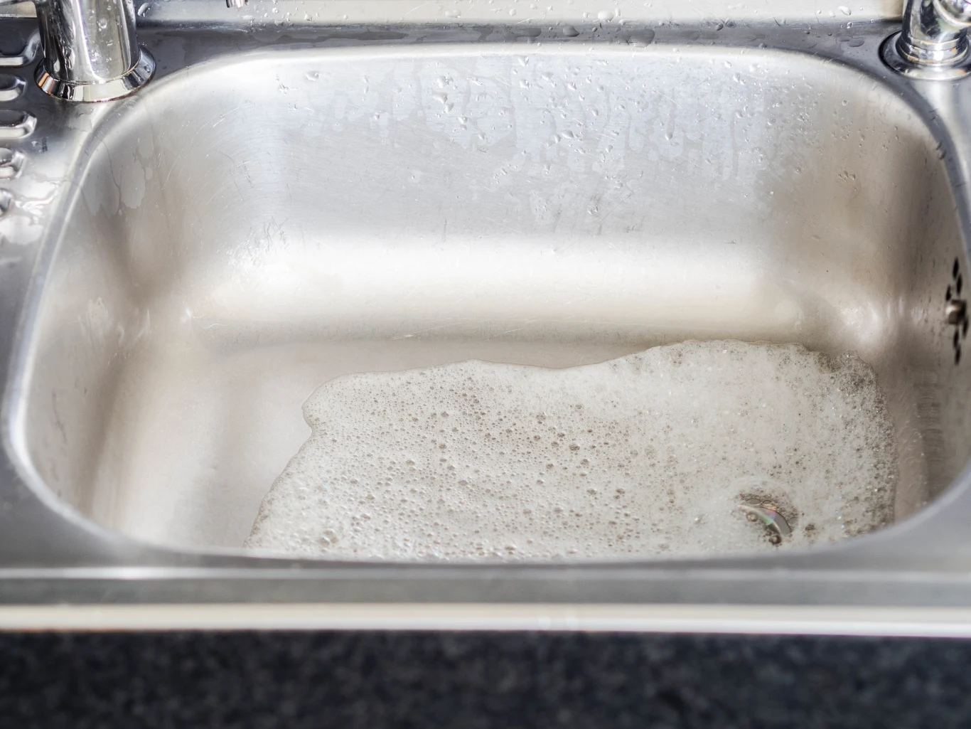 W wyniku połączenia sody i octu powstanie reakcja chemiczna - wytworzy się piana. To zapobiegnie nieprzyjemnym zapachom ze zlewu Stalowy zlew wypełniony częściowo wodą z pianą, z widoczną powierzchnią wilgotną wokół krawędzi oraz kranami po bokach.