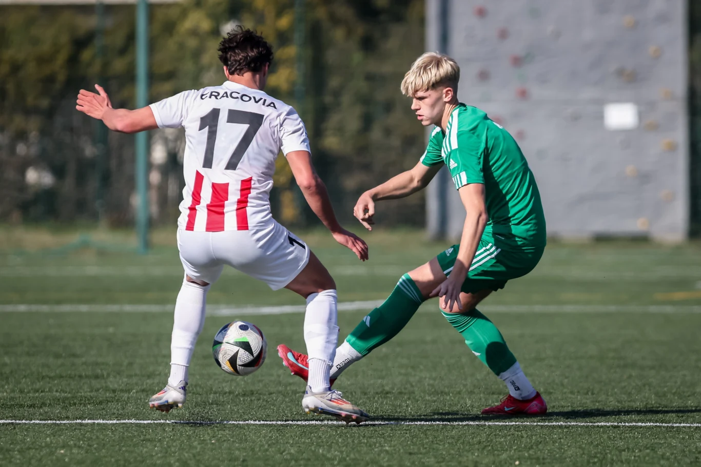 Dwóch piłkarzy rywalizujących o piłkę podczas meczu na trawie, jeden ubrany w biały strój z czerwonymi paskami i numerem 17, drugi w zielonym stroju sportowym.