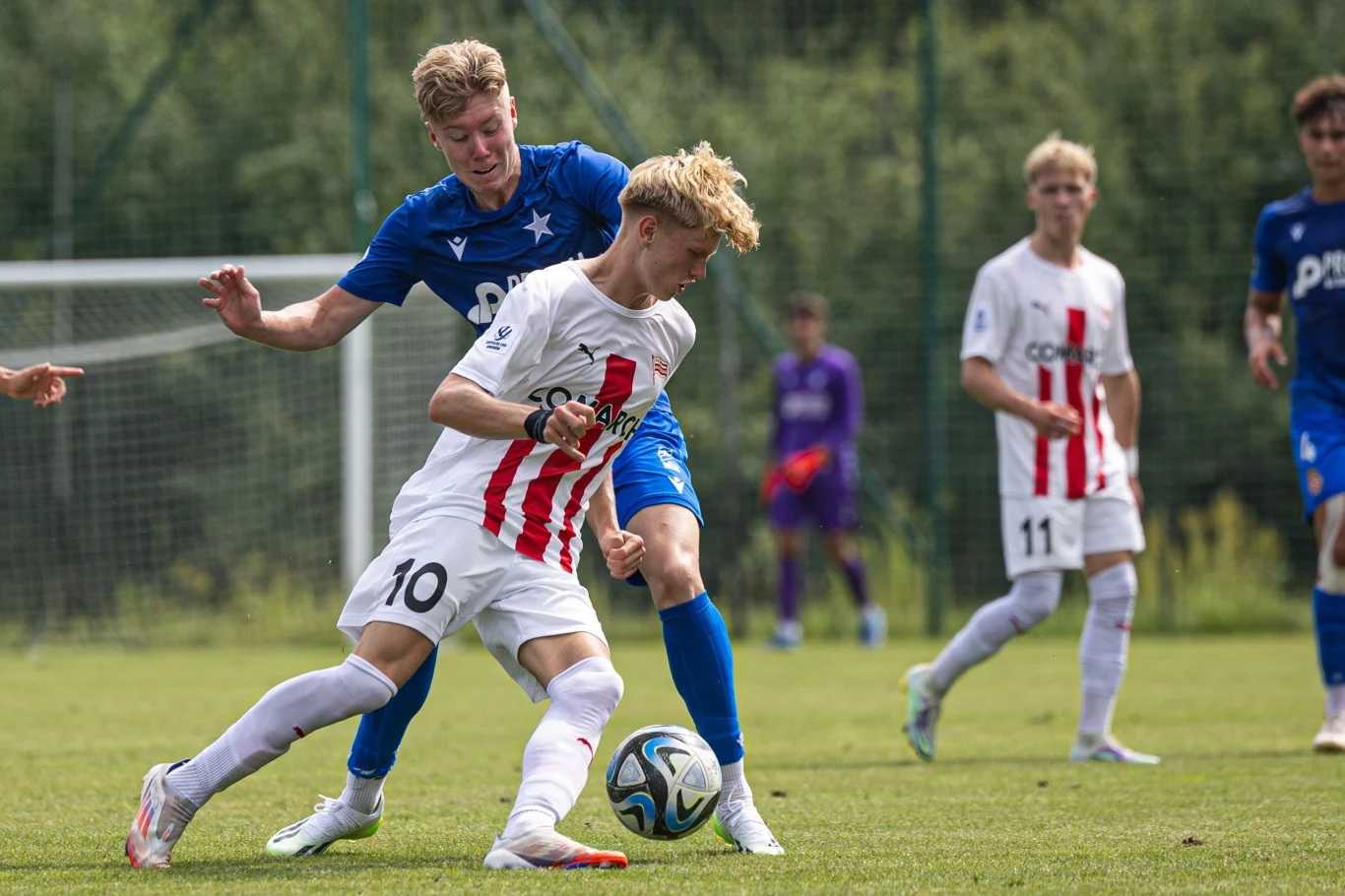 Dwóch młodych piłkarzy w trakcie rywalizacji o piłkę podczas meczu, jeden w białym stroju z czerwonymi paskami z numerem 10, drugi w niebieskiej koszulce z numerem 8, w tle widoczni pozostali zawodnicy oraz bramkarz.
