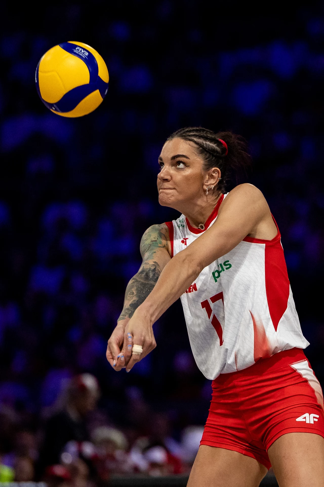 Malwina Smarzek Zawodniczka siatkówki w trakcie odbioru piłki, skupiona na piłce, ubrana w strój reprezentacyjny, dynamiczne ujęcie ruchu z wyraźnie widoczną piłką unoszącą się nad jej ramieniem, w tle rozmazana publiczność.