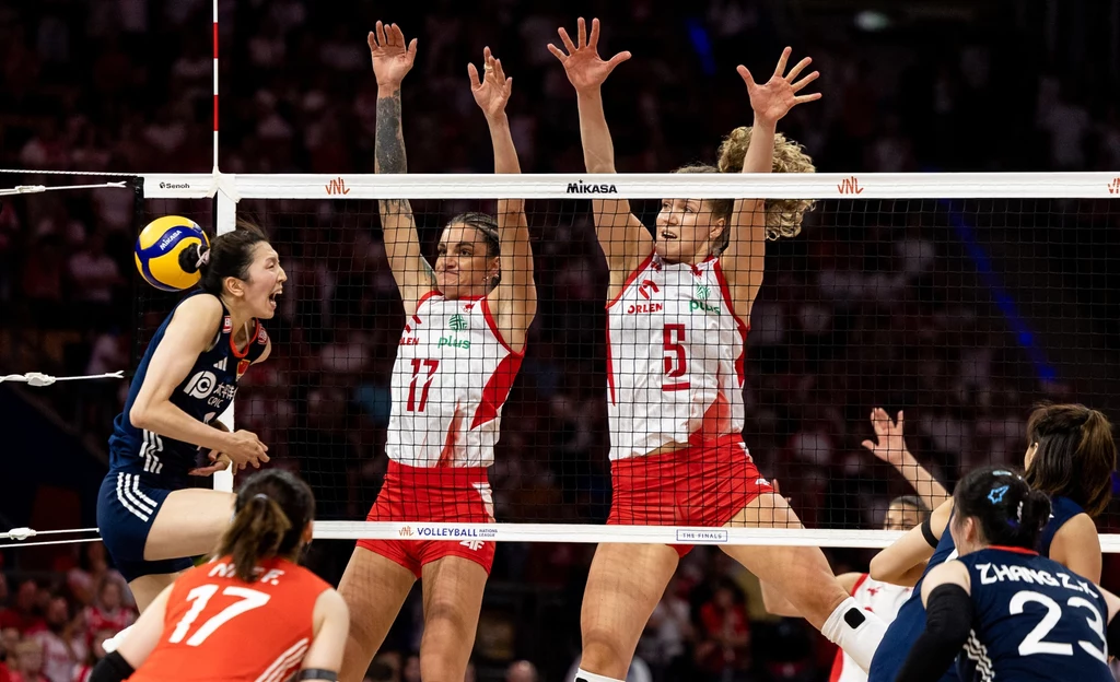 Malwina Smarzek w bloku (pierwsza od lewej) i Agnieszka Korneluk Siatkarki w trakcie dynamicznej akcji przy siatce, dwie zawodniczki w biało-czerwonych strojach wykonują blok, naprzeciwko nich zawodniczka w granatowym stroju uderza piłkę, pozostałe siatkarki skupione na przebiegu akcji.