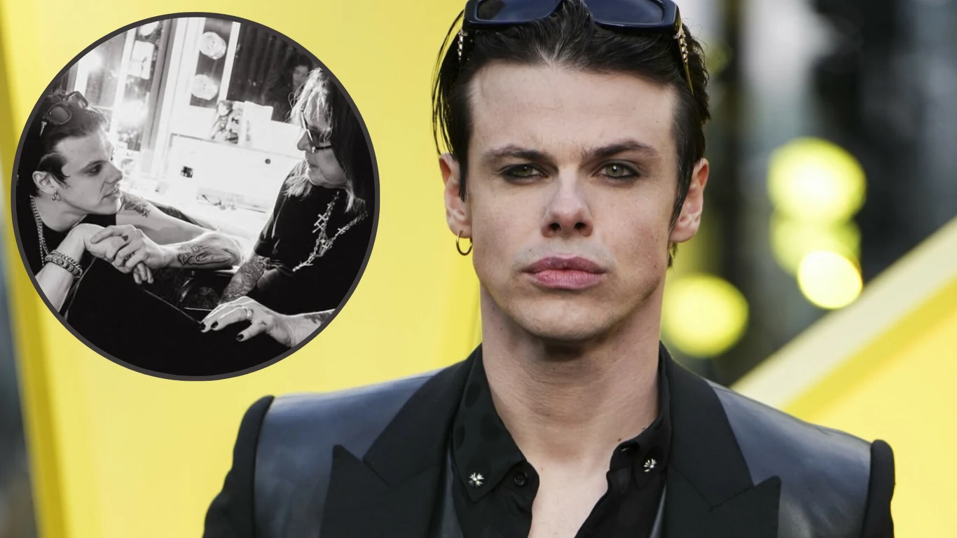 Yungblud Młody mężczyzna z ciemnymi, zaczesanymi do tyłu włosami, w czarnej marynarce i z okularami na głowie, stoi na tle żółtej struktury; w lewym górnym rogu okrągłe czarno-białe zdjęcie pokazuje tego samego mężczyznę podczas rozmowy z kobietą przy toaletce.