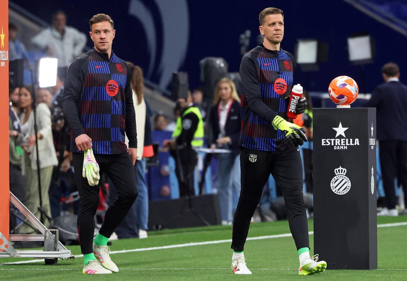 Marc-Andre ter Stegen i Wojciech Szczęsny Dwóch piłkarzy w strojach treningowych idzie po murawie stadionu, na pierwszym planie widoczny stół z piłką i logo sponsora, w tle liczni fotoreporterzy i kibice.