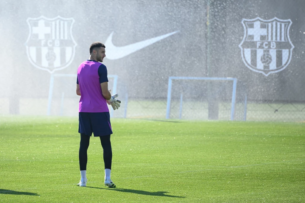 Joan Garcia w trakcie treningu Barcelony Piłkarz ubrany w różową kamizelkę treningową stoi na murawie boiska, w tle widoczne są dwa logotypy FC Barcelona oraz znak Nike, a także delikatna mgiełka wodna z systemu nawadniającego.