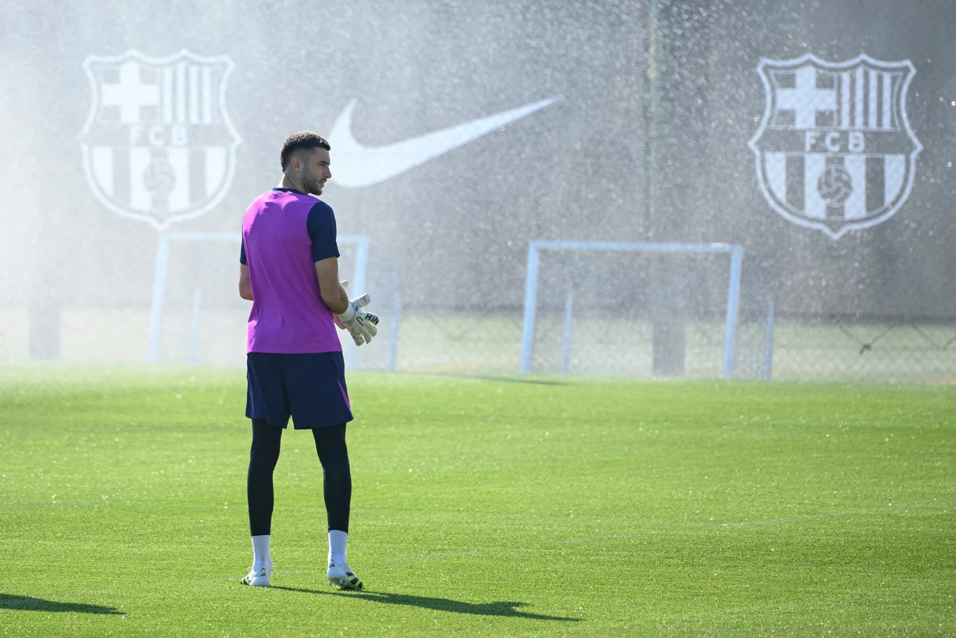 Piłkarz ubrany w różową kamizelkę treningową stoi na murawie boiska, w tle widoczne są dwa logotypy FC Barcelona oraz znak Nike, a także delikatna mgiełka wodna z systemu nawadniającego.