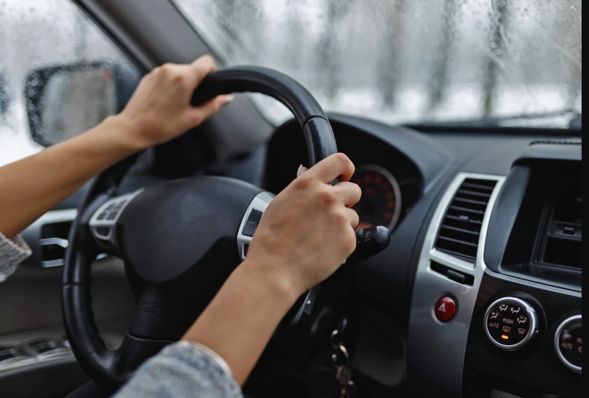 Drgania kierownicy po wymianie tarcz hamulcowych - przyczyny i rozwiązania. Ręce osoby trzymające kierownicę samochodu, widoczna część deski rozdzielczej z zegarami oraz lekko zamglona szybka przednia z kroplami deszczu na zewnątrz.