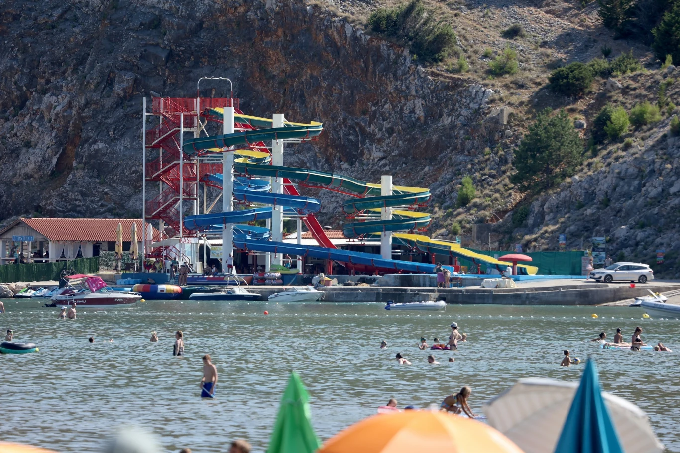 Park wodny Aquagan na chorwackiej wyspie Rab Wodny park rozrywki z kolorowymi zjeżdżalniami umieszczonymi przy brzegu, liczne osoby kąpią się w wodzie lub wypoczywają na plaży pod kolorowymi parasolami, w tle widoczny skalisty brzeg i roślinność.