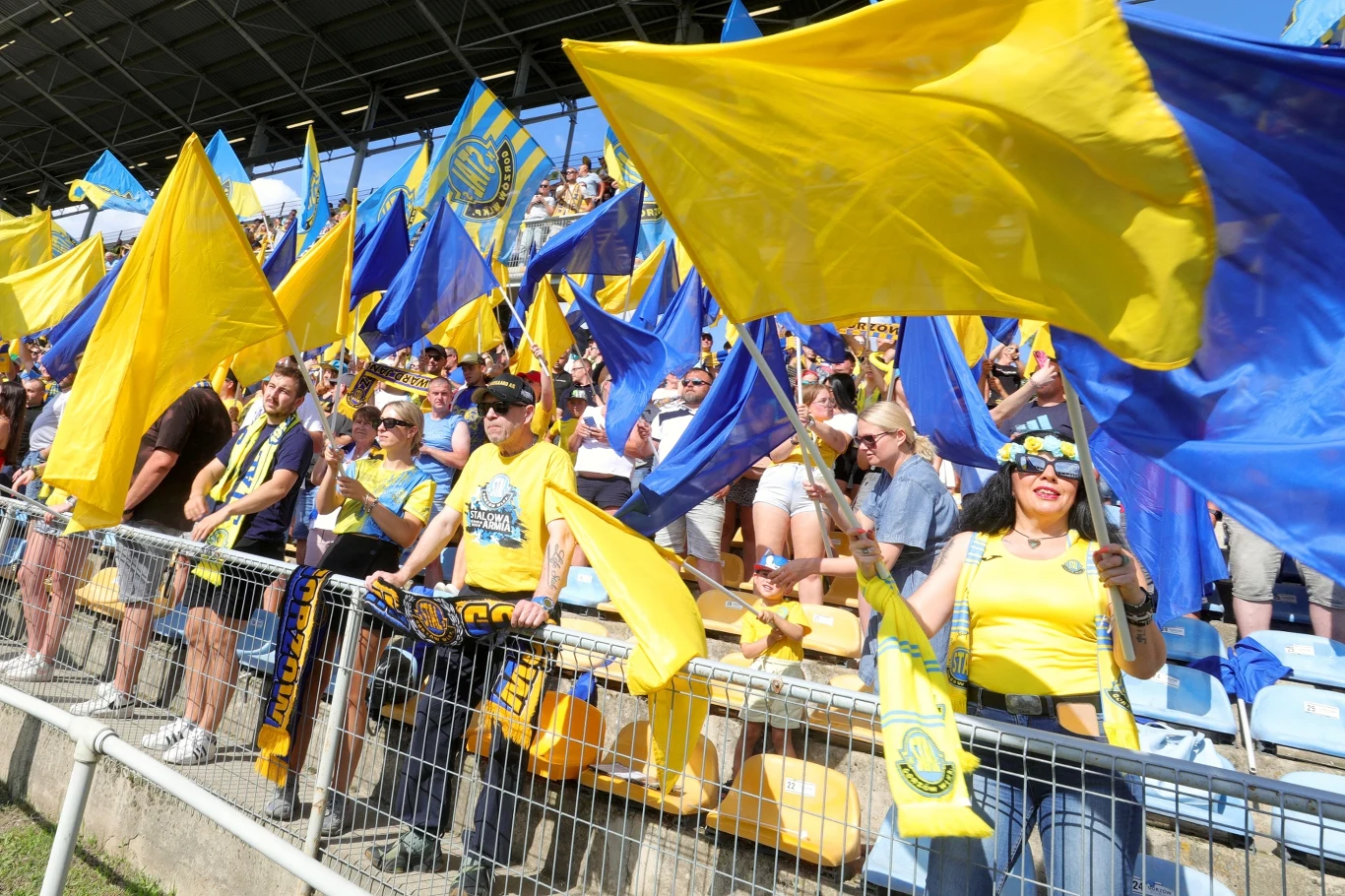 Kibice Stali Gorzów. Tłum kibiców ubrany w żółte i niebieskie stroje, machający flagami w tych samych kolorach na trybunach stadionu, atmosfera wsparcia i zaangażowania dla swojej drużyny.