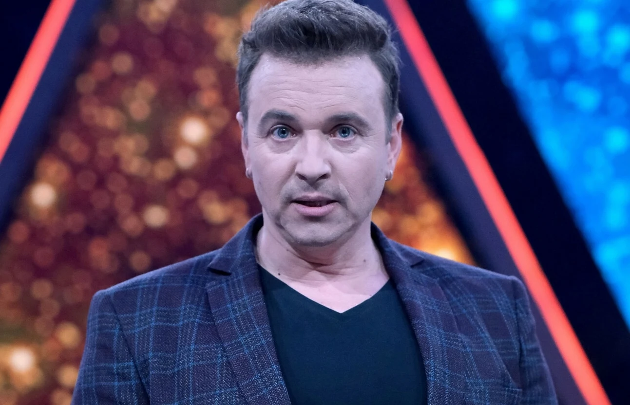 Radosław Liszewski trafił do szpitala Mężczyzna w marynarce w kratę i czarnej koszulce na tle kolorowych, rozmytych trójkątnych iluminacji scenicznych.