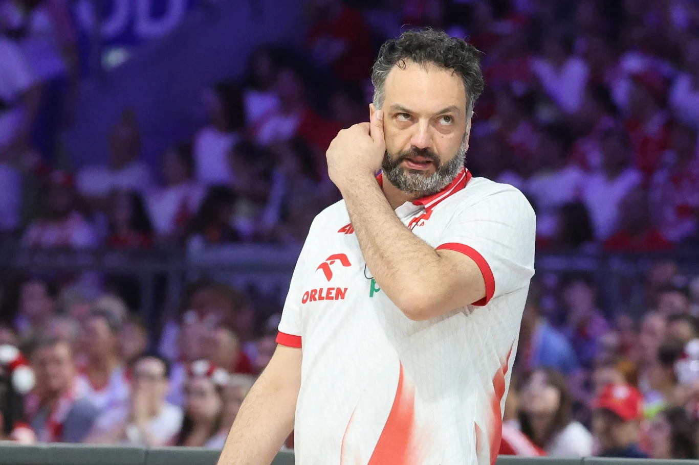 Mężczyzna ubrany w biało-czerwoną koszulkę z logo Orlen, będący trenerem bądź członkiem sztabu sportowego, stoi na hali sportowej podczas wydarzenia, w tle rozmyty tłum kibiców w biało-czerwonych barwach.