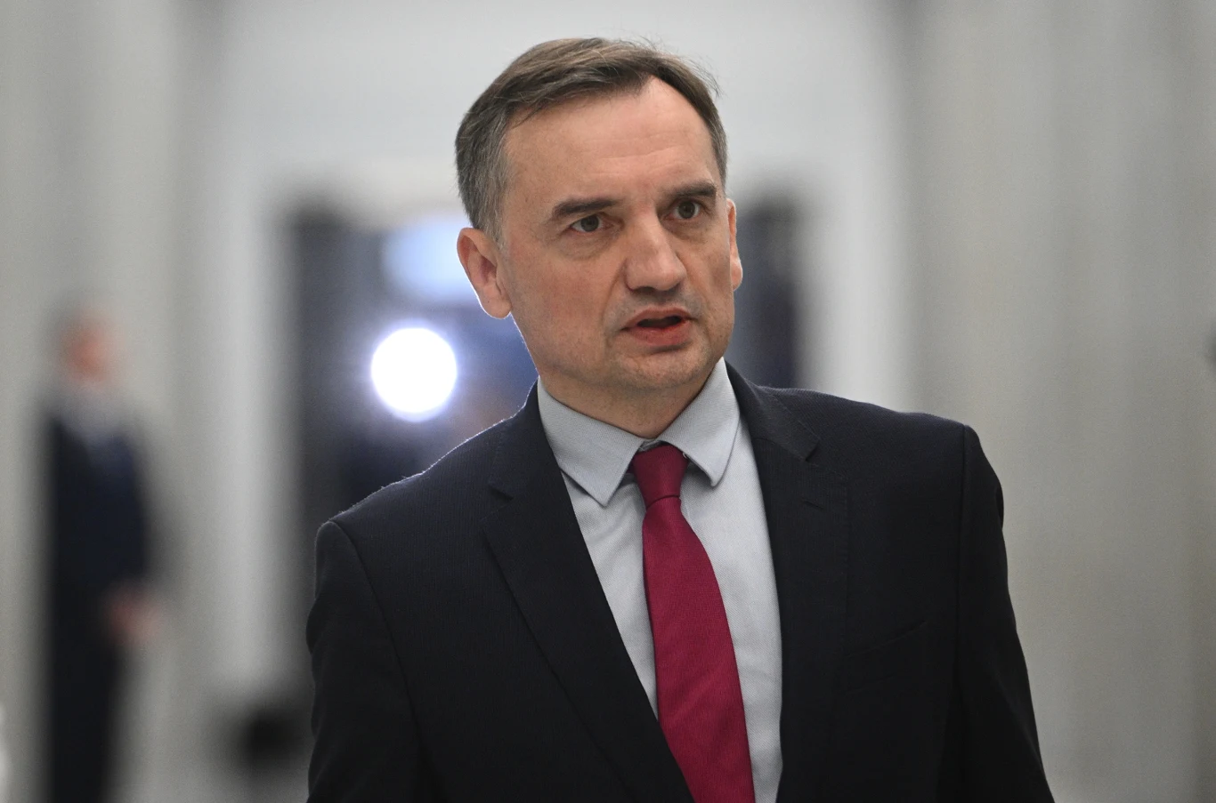 Zbigniew Ziobro Mężczyzna w garniturze i czerwonym krawacie stoi na korytarzu z rozmytym tłem, wyraz twarzy jest poważny, a światło pada na jego twarz z przodu.