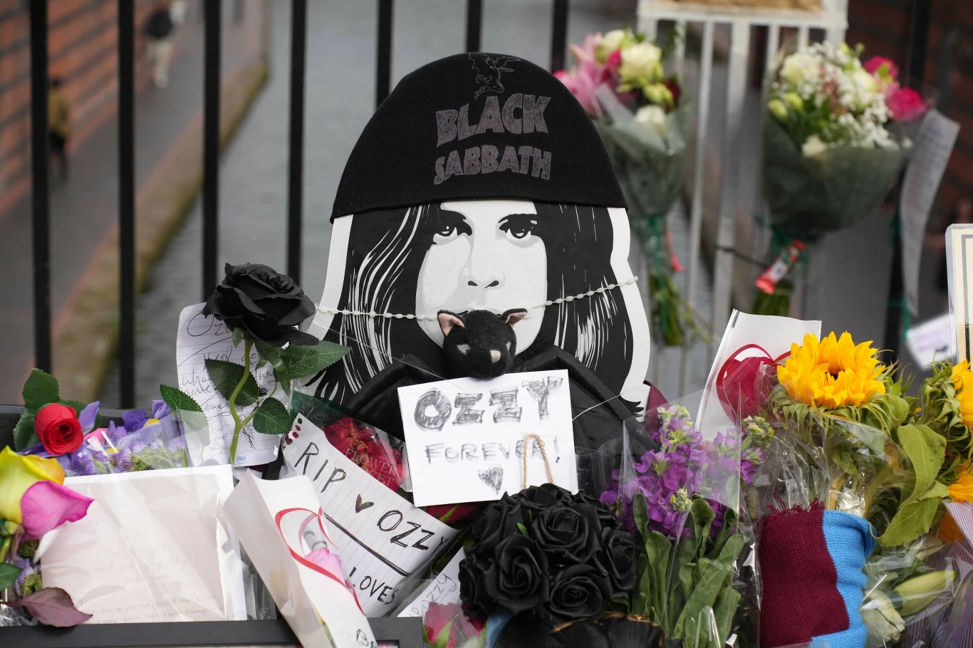 Wizerunek Ozzy'ego Osbourne'a na moście Black Sabbath w Birmingham utonął w kwiatach Portret osoby z napisem Black Sabbath otoczony kwiatami, kartkami z kondolencjami i symbolicznymi pamiątkami, w tle bukiety kwiatów ustawione przy metalowej barierce.