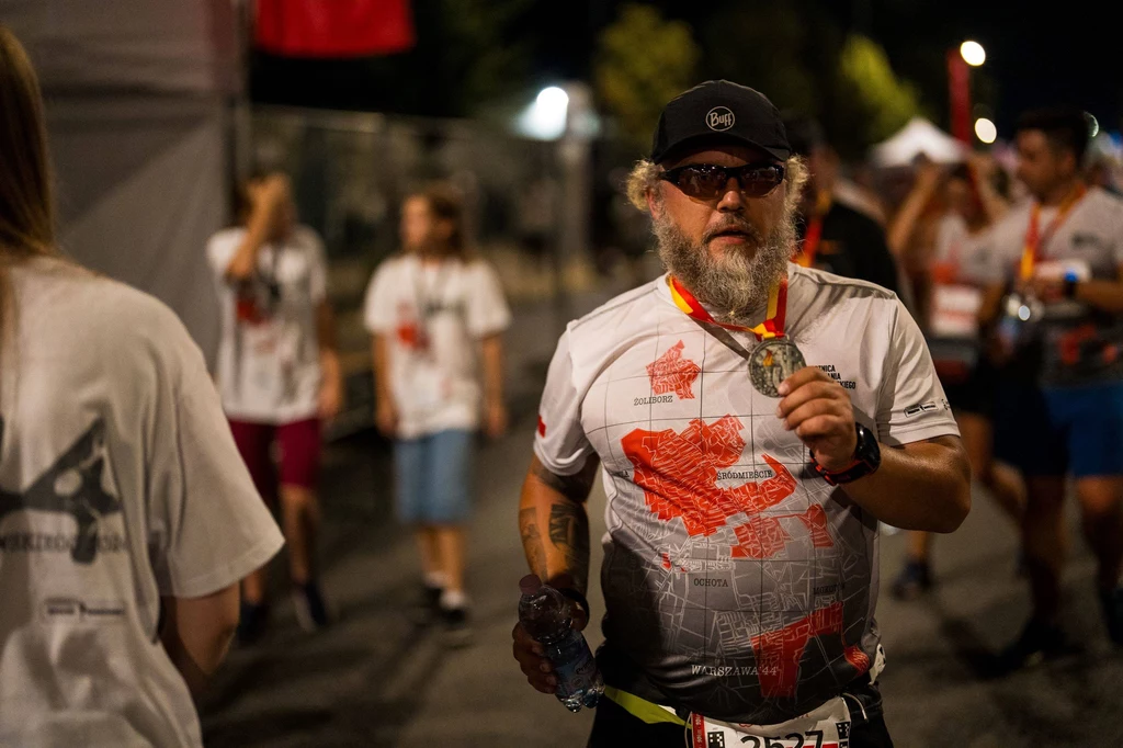 Bieg Powstania Warszawskiego Mężczyzna z brodą w okularach przeciwsłonecznych i czapce, ubrany w sportową koszulkę z mapą Polski, trzyma medal na szyi i butelkę wody, wokół niego inni uczestnicy biegu z medalami, atmosfera po zakończonej imprezie sportowej.