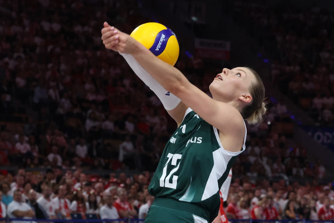 Siatkarka ubrana w zielony strój sportowy odbija piłkę podczas meczu siatkarskiego, w tle widoczna publiczność na trybunach.