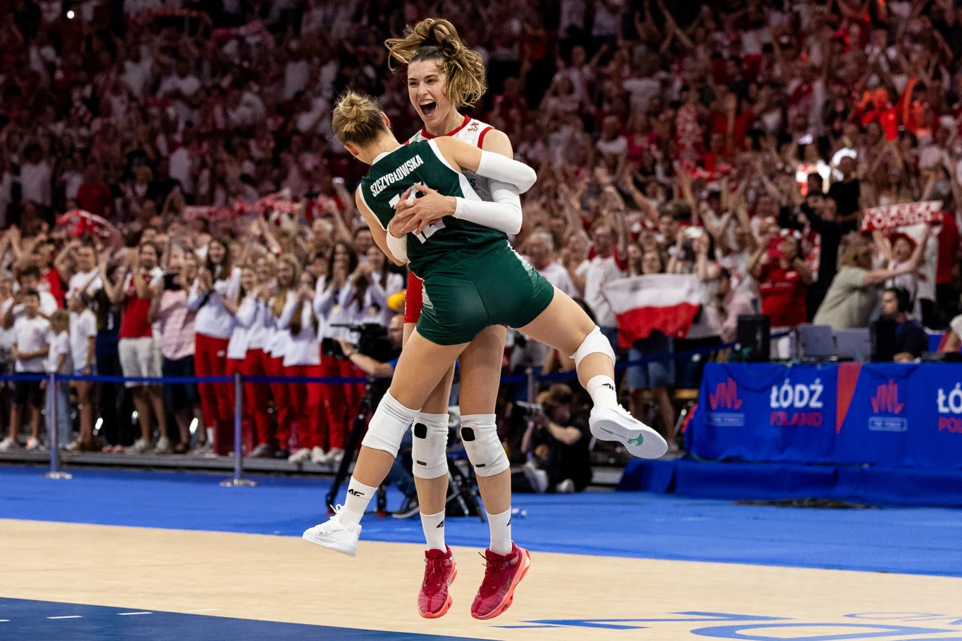 Dwie zawodniczki siatkówki w strojach reprezentacyjnych, wyskakujące w górę i obejmujące się w geście radości na boisku, w tle tłum kibiców ubranych w biało-czerwone barwy Polski z flagami.