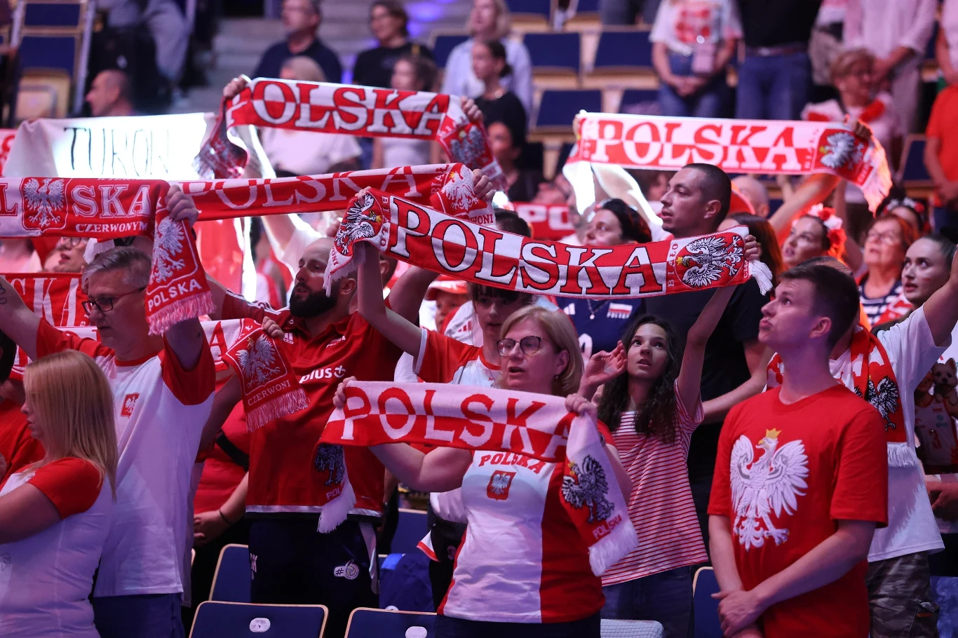 Grupa kibiców ubranych w barwy narodowe Polski z biało-czerwonymi szalikami z napisem 'Polska' podniesionymi nad głowami, wszyscy wyrażają wsparcie i entuzjazm podczas wydarzenia sportowego na trybunach.