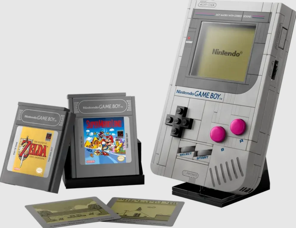 LEGO Game Boy Konsola Nintendo Game Boy w klasycznym szarym kolorze, obok widoczne trzy wymienne kartridże z grami, w tym The Legend of Zelda oraz Super Mario Land, wszystko ustawione na jasnym tle.
